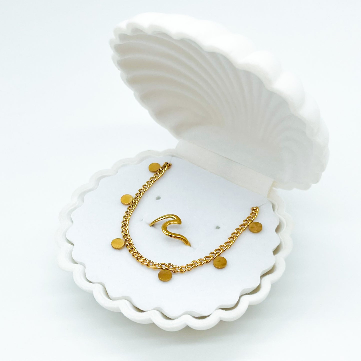 A set of jewelry including a wave ring and a mini coin necklace displayed on a white, shell-shaped dish.