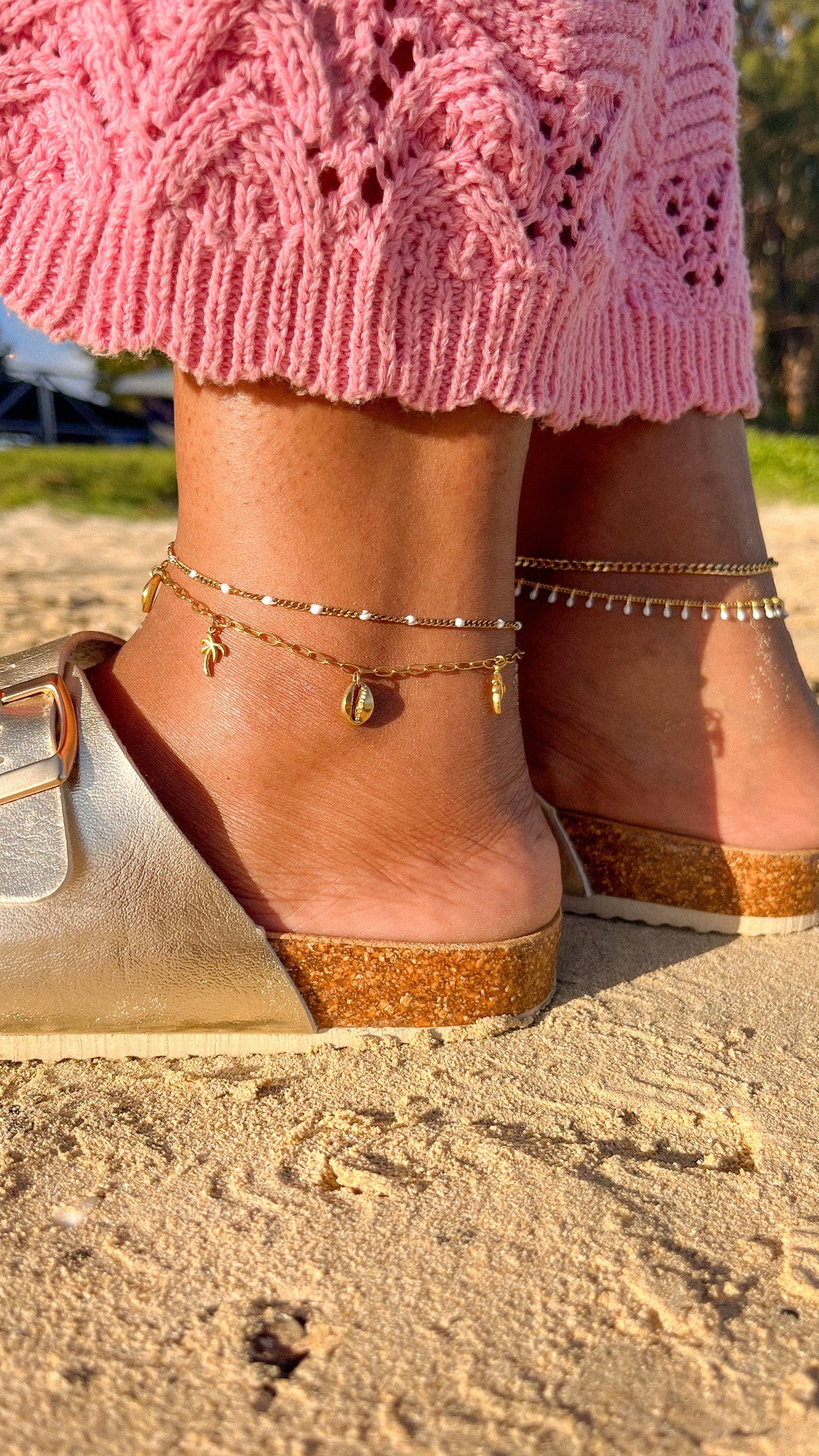 Close-up of feet wearing cork wedge sandals with gold anklets on a sandy surface.